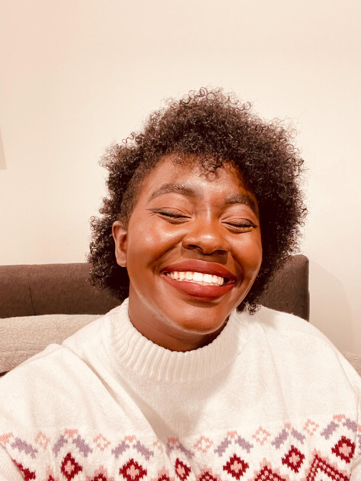 an image of Kammy, a dark-skinned Black nonbinary femme person with type 4B hair pulled to one side, smiling widely at the camera with their eyes closed. Kammy has on minimal brown eye makeup and burgundy lipstick. she is wearing a white sweater / jumper w